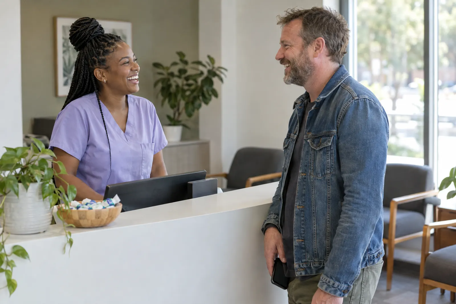 A patient coordinator in lilac scrubs laughing with a new patient in a denim jacket at a dental front desk.