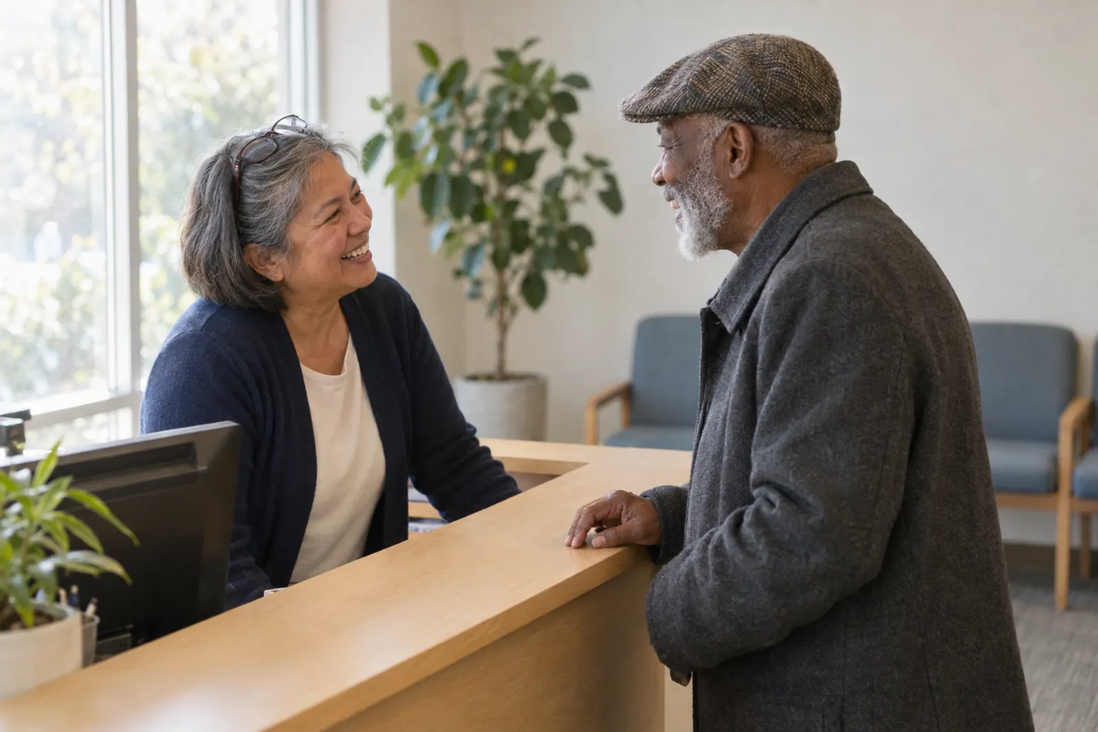 A front-desk coordinator laughing with a returning patient in a flat cap across a sunny clinic reception counter.