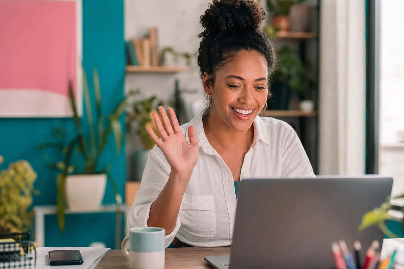 Smiling person waving during a laptop video call in a bright home office — illustrative photo