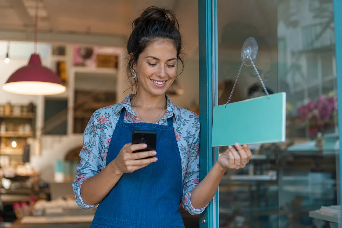 Small shop owner opening her storefront in the morning while checking her phone — illustrative photo