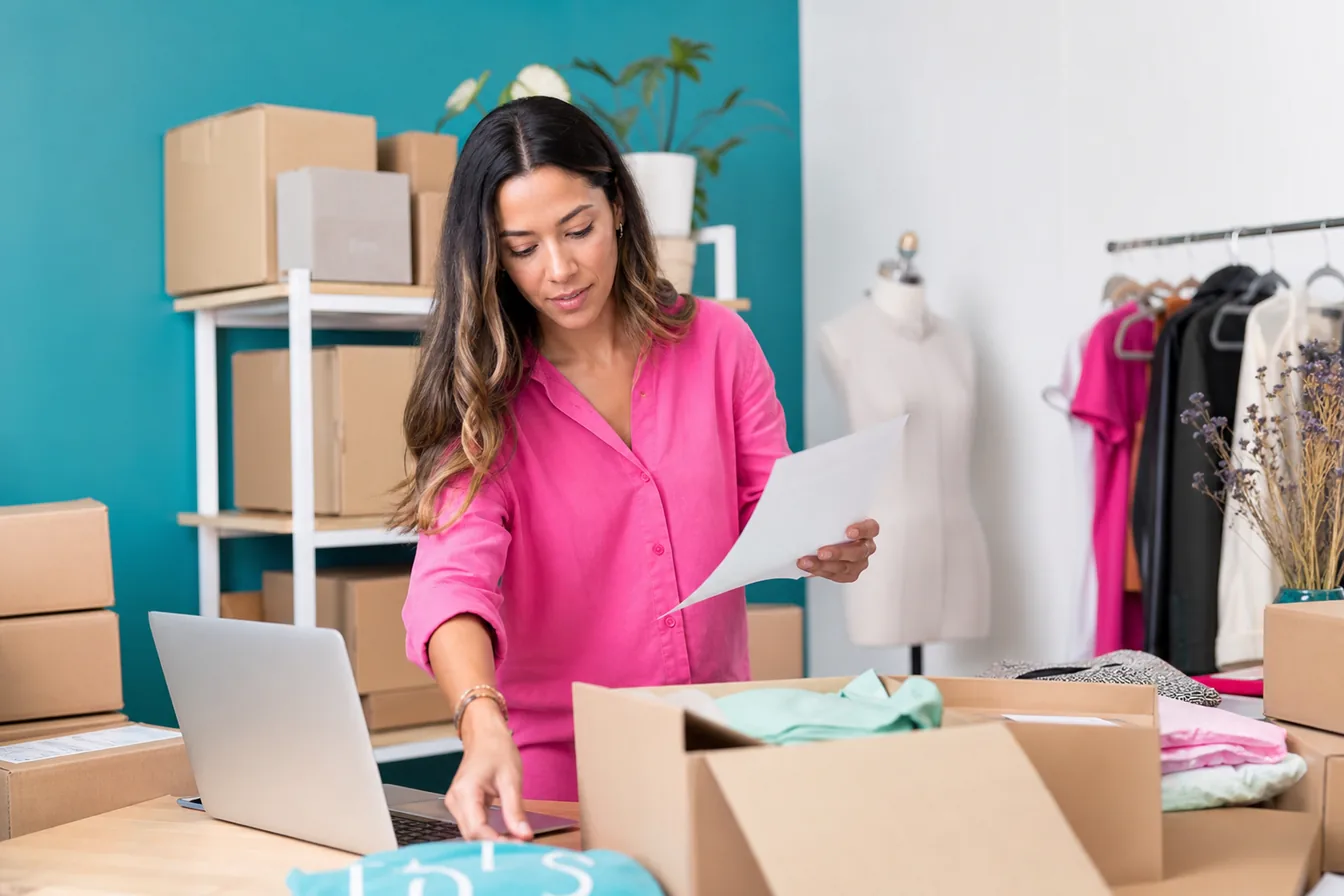Online-store owner among shipping boxes with a laptop on the packing table — illustrative photo